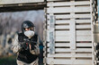 © qunica.com - A masked airsoft player in helmet and tactical vest aims a replica rifle behind a wooden barricade. Outdoor combat simulation scene with protective gear and focused stance.