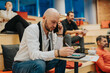 © qunica.com - A bald man in a white shirt writes notes while speaking on his mobile phone in a modern coworking space. In the background, several people sit on tiered seating, creating a casual work scene.
