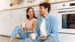 © Prostock-studio - A couple sits on the floor in a bright kitchen, holding mugs of coffee. They are smiling and engaged in a lively conversation while enjoying their time together in the morning light.