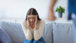 © Prostock-studio - A woman looks distressed while sitting on a couch with her hands on her head. A person stands nearby, suggesting a tense situation. The setting is a simple living room with plants.