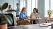 © Creative Spectrum - Two women collaborating with a laptop at a small round table in a modern, bright office setting while colleagues talk in the background