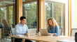 © Creative Spectrum - Two colleagues working on laptops at a wooden table in a modern office with large windows showing greenery outside, focused on their screens