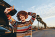 © SHOTPRIME STUDIO - Happy woman with curly hair wearing rainbow sweater taking selfie with smartphone near car outdoors, smiling brightly, sunny day, palm trees and clear sky background.