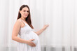 © New Africa - Beautiful pregnant woman touching her belly near curtains at home, space for text
