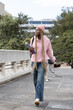 © wavebreak3 - Female student walking on campus bridge wearing pink hoodie beanie holding notes checking cell