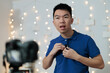 © fotolite - A young Asian man clips a microphone to his shirt while preparing to record in front of a camera in a home studio, ideal for content creation setup, behind the scenes, or UGC production concepts.