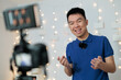© fotolite - A smiling Asian young man wearing a microphone records himself on camera in front of fairy lights in a home studio, ideal for content creation, social media influencer, or UGC marketing concepts.