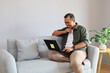 © ArvStd - A handsome man relaxes comfortably at home, sitting on the sofa, focused on his laptop with a relaxed smile.