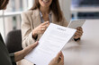 © fizkes - Close up image of female holding and reading printed contract, while manager or business partner smiles and engages in conversation, holding digital tablet. Buying and selling services, making deal