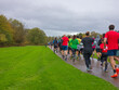 © Paul - A large group of runners viewed from behind running along a glistening wet tarmac park path on an overcast autumn morning, with vivid green grass verges and golden autumn woodland lining the route