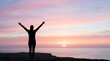 © Luka - Silhouette of a woman with raised arms standing on a cliff overlooking the ocean at sunset. Freedom, success and mindfulness concept.