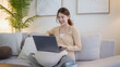 © wattana - Young woman smiling while using laptop on cozy living room sofa.