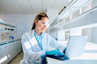 © we.bond.creations - Young female scientist wearing lab coat, safety glasses, and gloves, working on a laptop in a modern laboratory