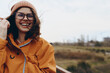 © SHOTPRIME STUDIO - Smiling woman in orange jacket and brown beanie enjoys outdoor lifestyle on a windy day. She wears glasses and has loose hair with a blurred natural landscape in the background.