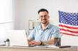 © Pixel-Shot - Handsome young happy man with laptop and flag of USA working at table in office