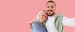 © Pixel-Shot - Young man hugging his mother and taking selfie on pink background