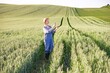 © sofiko14 - A woman in overalls and a straw hat examines a wheat stalk while standing in a vast, green field with rolling hills in the background