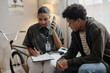 © Seventyfour - Black female young adult social worker talking on smartphone while writing on clipboard, sitting beside Black male teenager in casual clothing during counseling session indoors