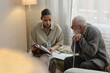 © Seventyfour - Young adult Black woman social worker discussing documents with senior Caucasian man using cane, both sitting closely together in home setting, focusing on paperwork interaction