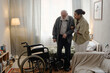 © Seventyfour - Black woman social worker assisting senior Caucasian man with disability standing up from bed in home setting, wheelchair positioned nearby, supportive interaction evident