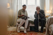 © Seventyfour - Young adult Black woman social worker sitting on bed holding clipboard, talking with senior Caucasian man with beard using cane in residential room during consultation session