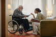 © Seventyfour - Black woman social worker assisting senior Caucasian man in wheelchair reading brochure in home setting, both engaged in caregiving interaction, showing attentive support