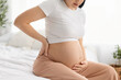 © Anastasiya - Close up of pregnant woman holding belly and touching lower back while sitting on bed at home. Concept of pregnancy back pain, prenatal health problems and maternal wellbeing
