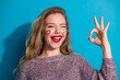 © deagreez - Cheerful young woman with face art bright lipstick smiling against blue background wearing a cozy sweater and playful hand gesture
