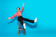 © deagreez - Cheerful mature businesswoman sits on a wooden stool kicking her legs with flair against a bright blue background smiling and enjoying a playful fashion moment