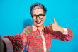 © deagreez - Confident mature businesswoman smiling with thumbs up wearing a colorful zigzag jacket blue background taking selfie and promoting positivity and style
