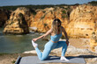 © Angelov - A woman in athletic clothes stretches on a yoga mat on the beach. A beautiful woman meditates, relaxes, or does a workout by the sea. Concepts of yoga, sports, and enjoyment.