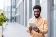 © Liubomir - Young handsome man wearing an earth tone shirt smiling while holding and interacting with a modern digital tablet, connecting to technology and managing professional work in an urban environment