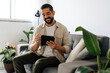 © kleberpicui - Man sitting comfortably on a sofa using a tablet in a bright living room, representing leisure, technology use at home and a relaxed modern lifestyle.