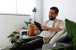 © kleberpicui - Man sitting comfortably on a sofa using a tablet in a bright living room, representing leisure, technology use at home and a relaxed modern lifestyle.