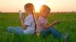 © Victoriia - Children play online games with their phones in hand in nature. Kids on green lawn park use smartphones. Boy, girl communicate via smartphones. Kids play on green grass using modern technology