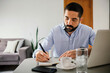 © kleberpicui - Man sitting at a table writing notes while using a laptop at home, representing remote work, productivity, planning and modern professional lifestyle.