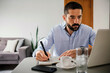 © kleberpicui - Man sitting at a table writing notes while using a laptop at home, representing remote work, productivity, planning and modern professional lifestyle.