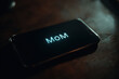 © britaseifert - A smartphone resting on a dark wooden table,incoming call labeled 'Mom', glowing brightly on the screen