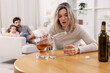 © New Africa - Alcohol addiction. Woman drinking whiskey at wooden table while her scared husband and daughter sitting on sofa indoors, selective focus