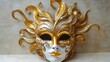 © RATAN - A gold and white Venetian mask with intricate designs and swirling patterns sits on a surface against a neutral background.