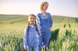 © sofiko14 - A woman in overalls and a straw hat stands with a young girl in a wheat field at sunset, both wearing denim shirts
