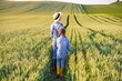 © sofiko14 - A woman and a young girl walk through a lush green wheat field, with rolling hills and cultivated land in the background under a bright sky