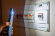 © Nanci - Person holding smartphone flashlight to illuminate a tripped electrical fuse box during a home power outage, highlighting emergency troubleshooting and domestic electrical issues
