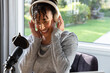 © wavebreak3 - African American woman adjusting headset in gray sweater, smiling at window with mic and pop filter