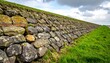 © Gani - Stone wall slopes diagonally, covered in moss and grass, reaching towards the sky under a cloudy blue sky