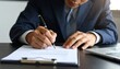 © gelas - Suited man signs paperwork on a desk with a pen, notebook, and natural light from behind