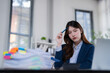 © MINAE - Stressed young business woman sitting at a desk in an office, holding a pen to her head, surrounded by overflowing documents, feeling overwhelmed and experiencing burnout from work