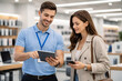 © ольга  - Modern office has shelves with books and computers. Man in a blue polo shirt holds a tablet. He wears a lanyard and smiles while showing it. Woman in a beige blazer examines the screen