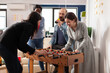 © DC Studio - Office workmates playing foosball table game together after hours, enjoying alcoholic drinks and pizza. Man and women doing party celebration and having fun with match after work.