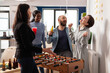 © DC Studio - Cheerful woman winning foosball game competition at office party after work, having fun with coworkers. Businesspeople playing soccer match at table, eating snacks and drinking beer.